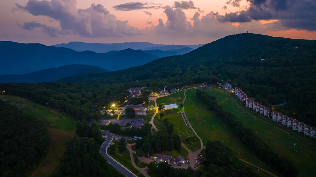Aerial view at dusk of the mountain village at Wintergreen: lit lodges and roads, a grassy slope lined with condominium buildings, and blue ridges under a cloudy sunset.